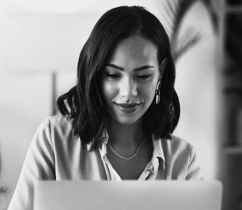 A woman with shoulder-length dark hair smiles while working on a laptop in a bright, indoor setting.