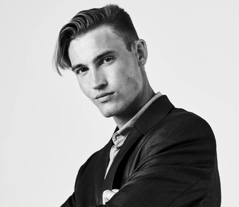 A young man with slicked-back hair and a confident expression poses in a suit against a plain background.