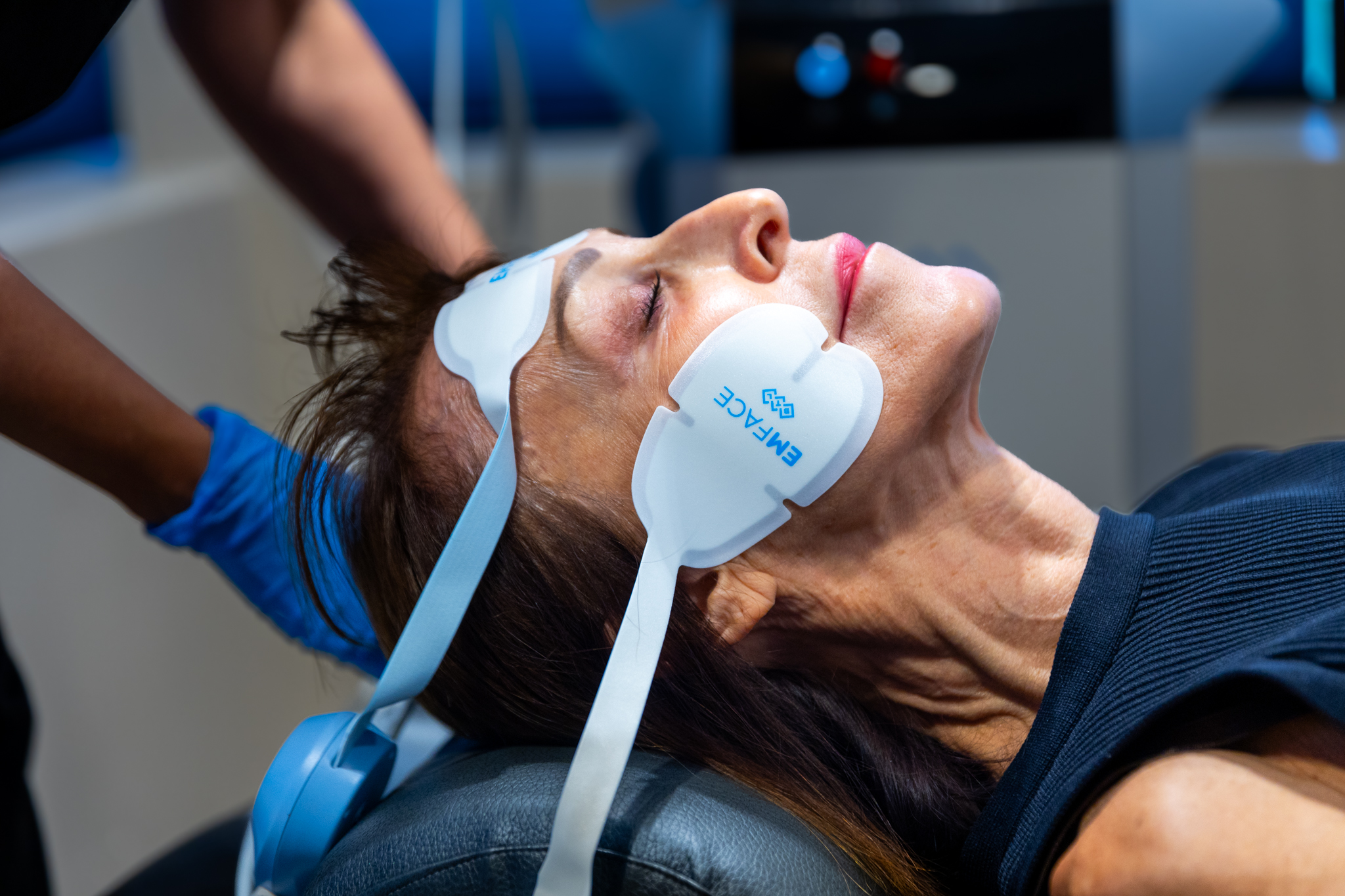 An older woman receiving EMFACE treatment, a non-invasive facial therapy using a specialized headgear device in a medical setting.