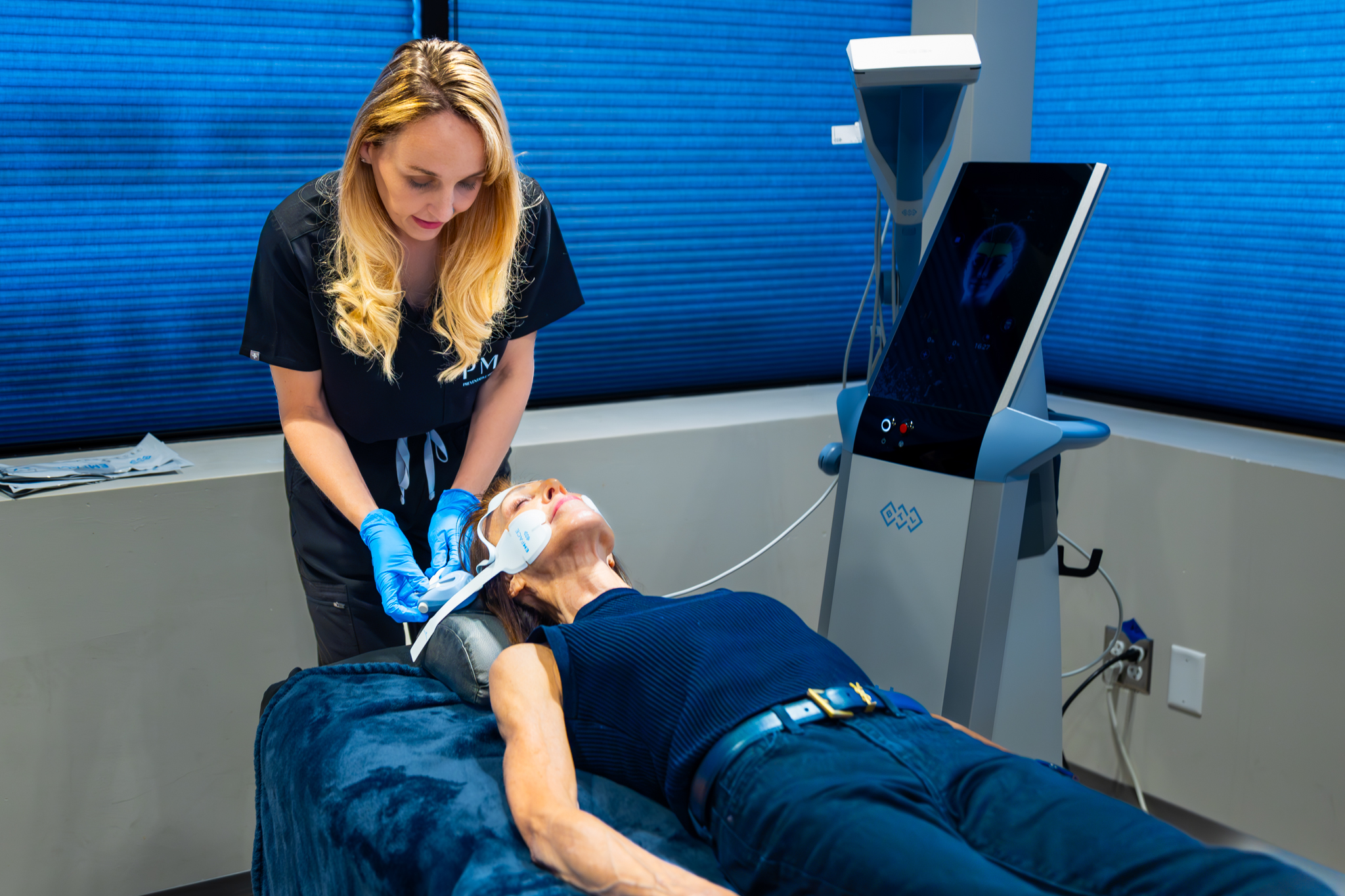 A female medical practitioner administers an EMFACE treatment to a reclining male patient in a modern clinic, with a sophisticated device screen visible in the background.