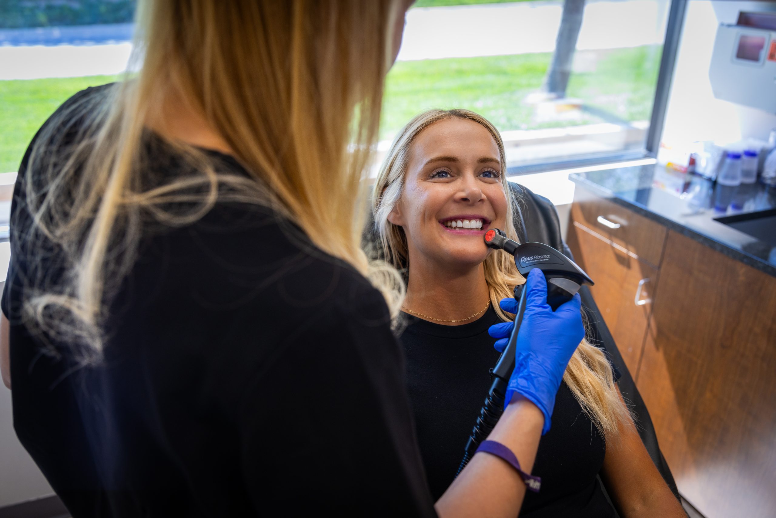 A smiling woman receiving Opus Plasma treatment on her face from a healthcare professional in a clinical setting, enhancing her skin texture and appearance.