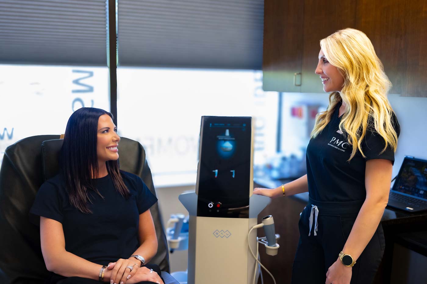 A patient smiles while sitting in a treatment chair, conversing with a clinician who is operating an Exion machine in a modern medical office.