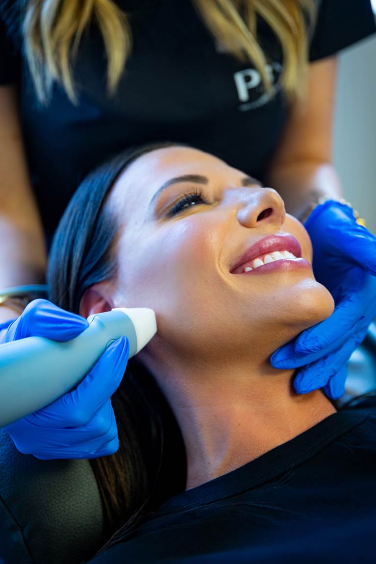 A smiling woman receives a non-invasive Exion treatment on her neck, with a practitioner applying a handheld device in a clinical setting.