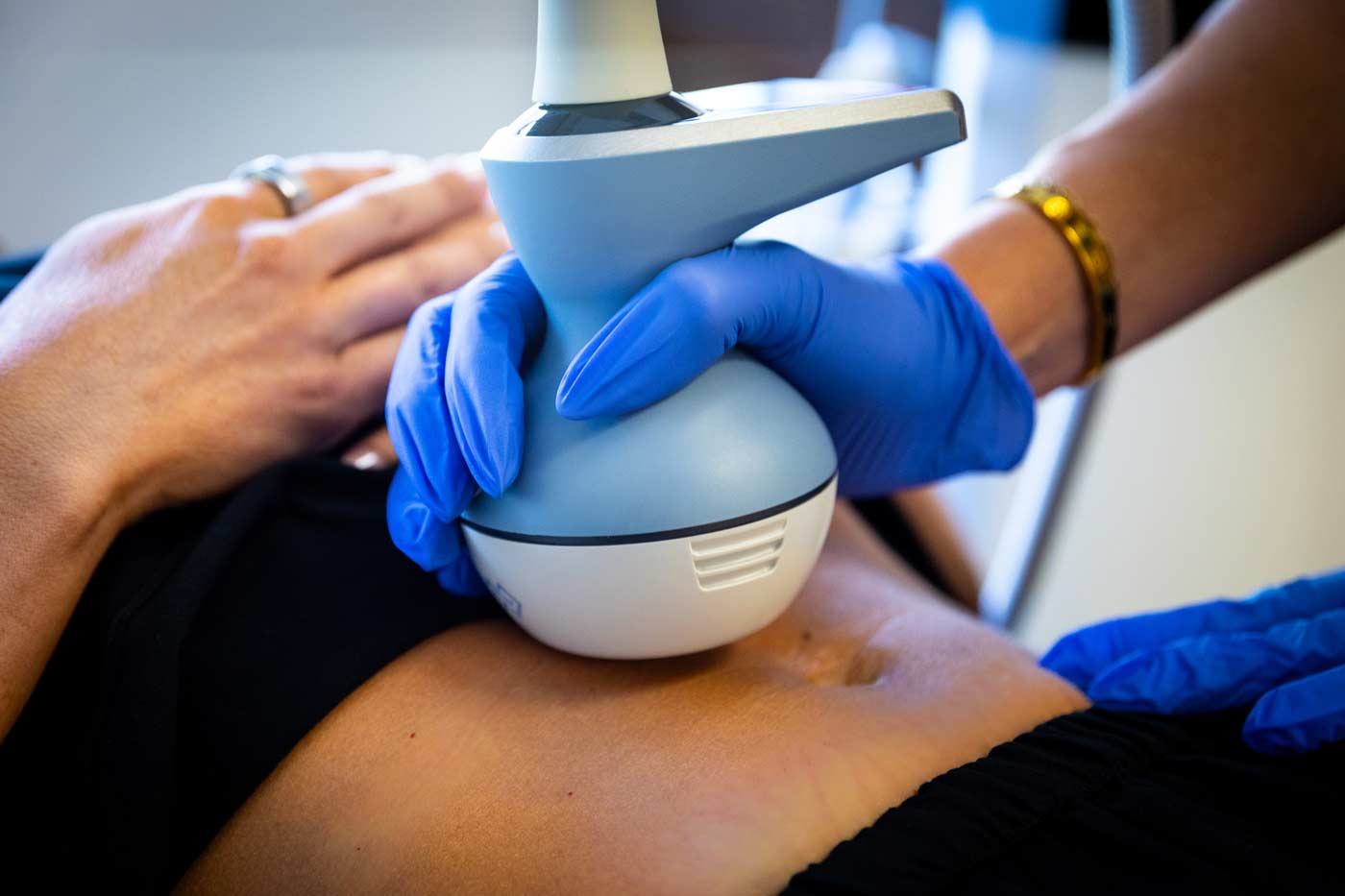 Close-up of a practitioner's hands wearing blue gloves, using an Exion device on a patient's abdomen in a clinical environment.