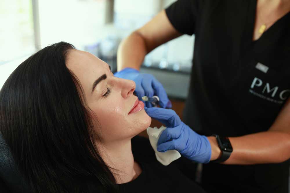 A patient relaxes with her eyes closed while receiving a filler treatment, as a medical professional carefully applies an injection to her cheek in a well-equipped clinic.