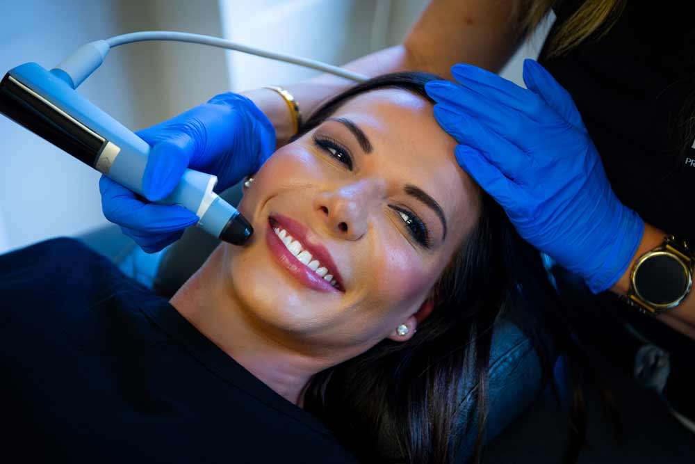 A content patient receives a facial treatment as a clinician, wearing blue gloves, expertly handles a modern device along the patient's jawline in a clinical setting.