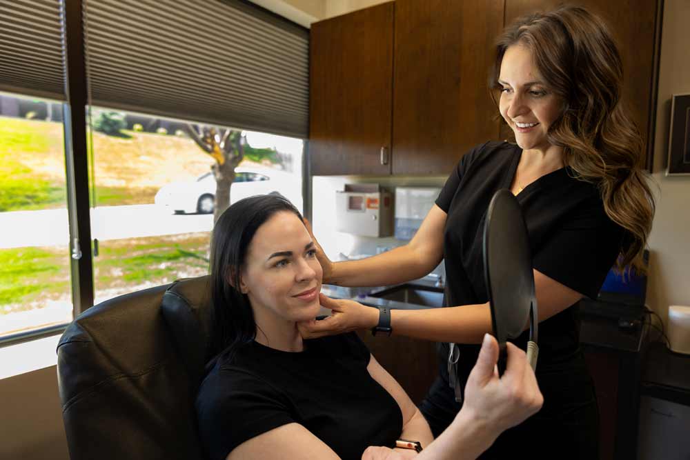 A patient examines her facial results in a handheld mirror, smiling as a medical professional gently touches her chin after a treatment in a well-lit clinic.