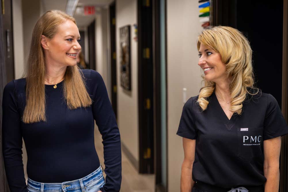 Two women, a medical professional in a dark uniform and a patient in casual attire, share a friendly conversation in a clinic hallway, both smiling and looking at each other.