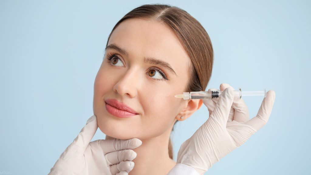 A woman with clear skin receiving a cosmetic injection near her eye from a professional wearing white gloves. (MODEL)